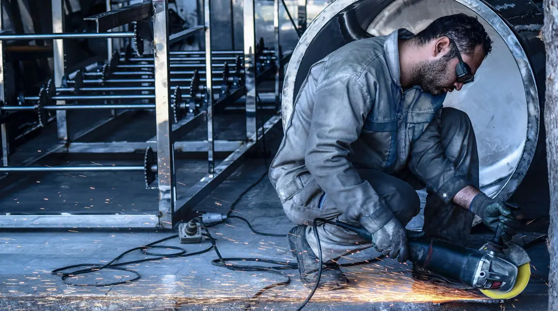 Metal fabricator grinding a large steel cylinder, sparks flying in an industrial shop