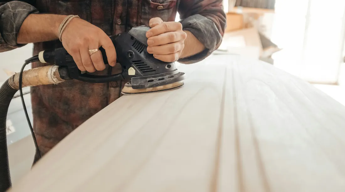 Carpenter using an orbital sander to smooth a wood plank in a sunlit workshop