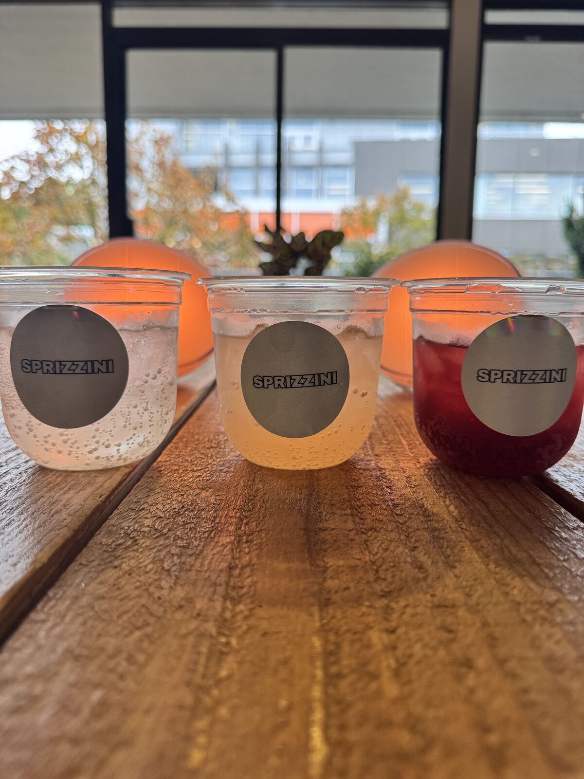 Three clear cups of Sprizzini on a long wooden table, each wearing a holographic Sprizzini sticker