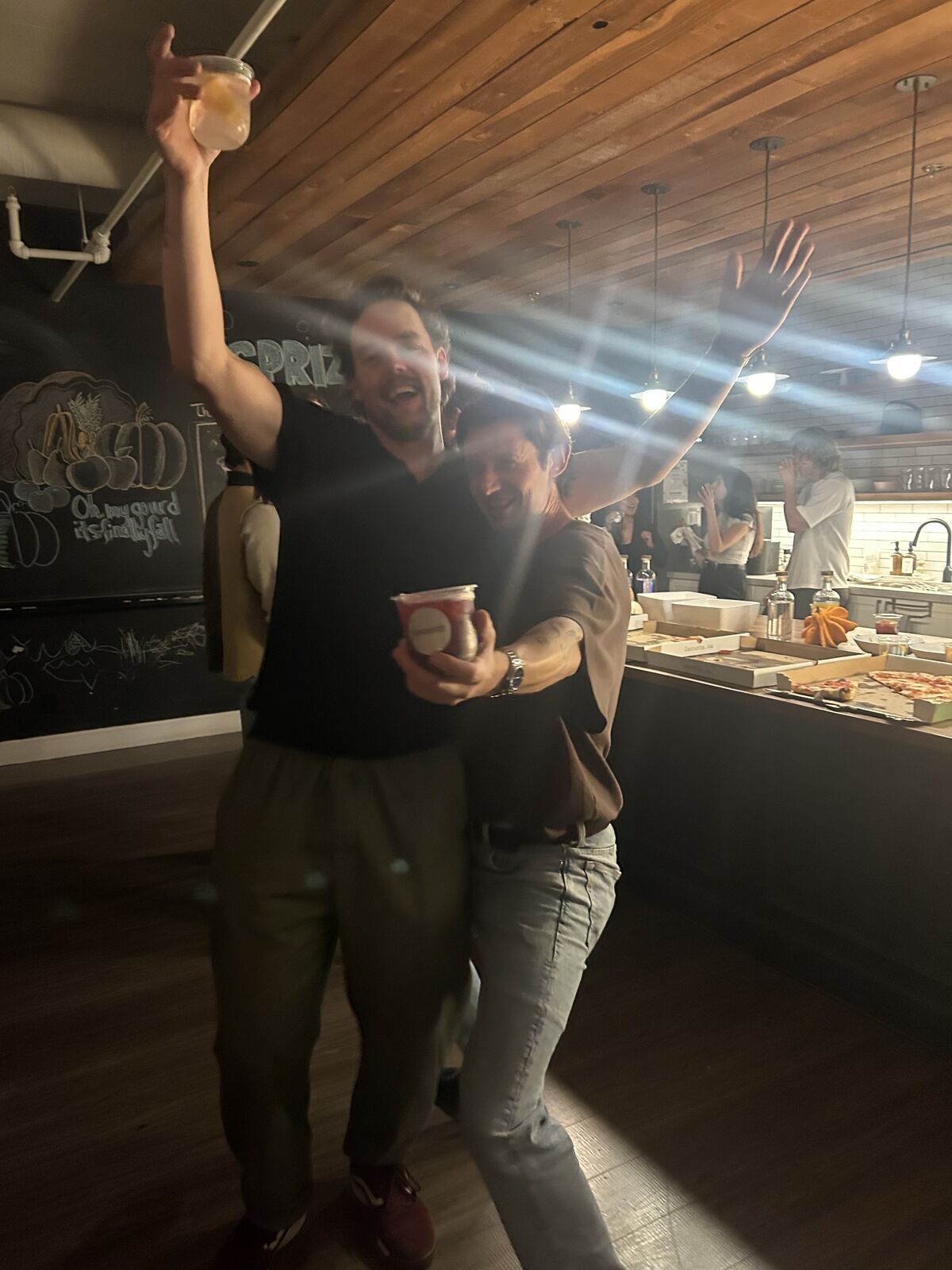 Two guests with arms raised, drinks in hand, in front of the chalkboard