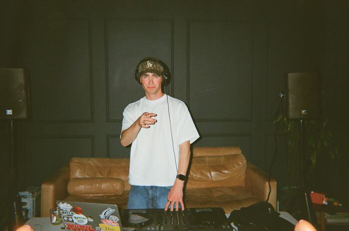 Liam in a white tee and camo cap pointing at the camera from behind the decks