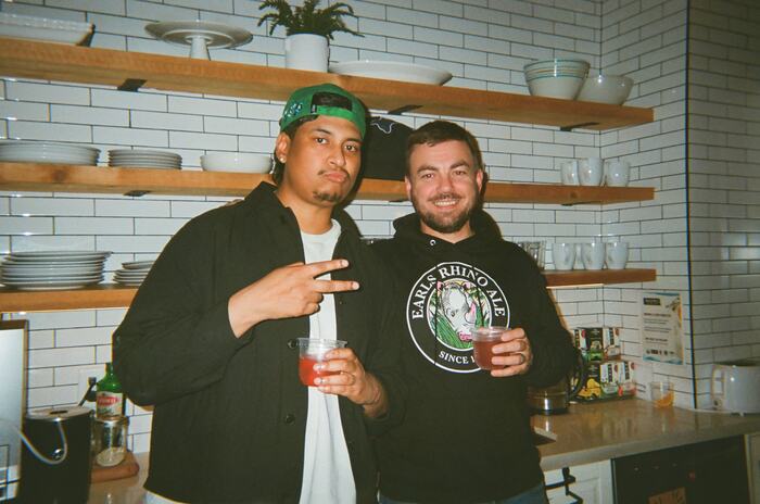 Two guests in the kitchen holding red drinks, one throwing a peace sign