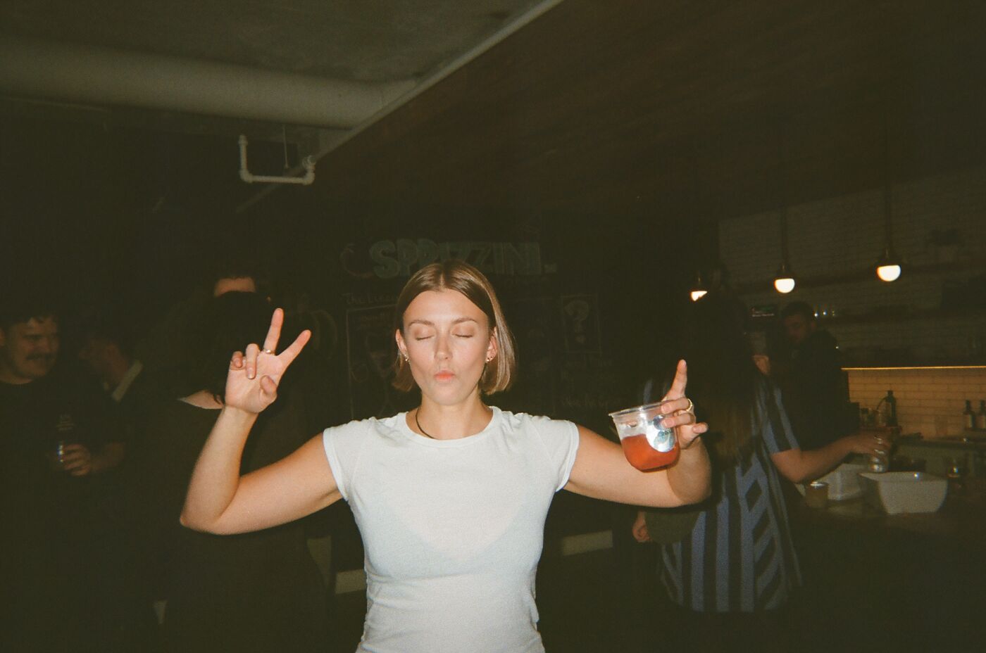 A guest in a white tee dancing with her eyes closed, arms up, a red drink in one hand
