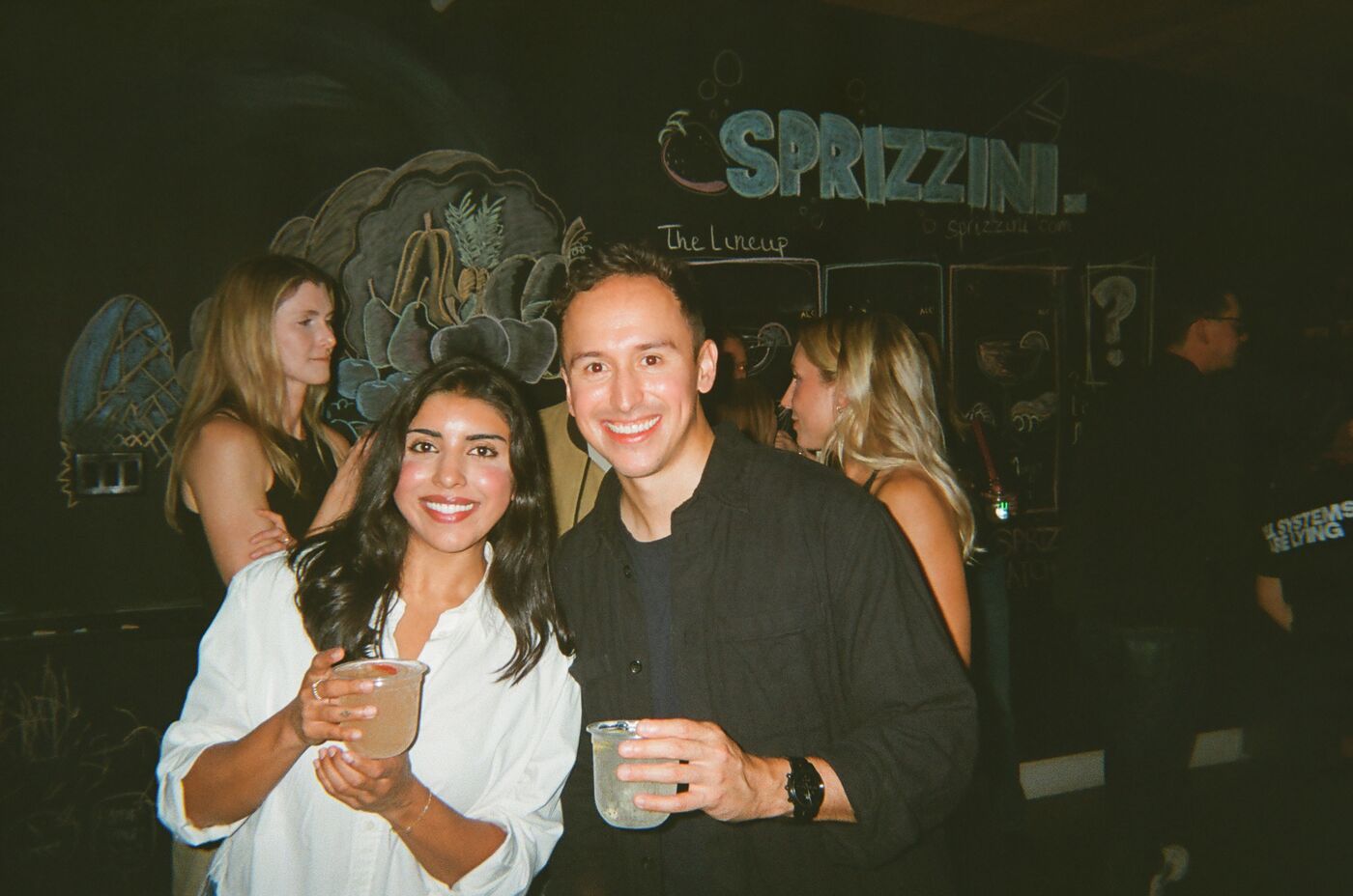 Two guests holding pale spritzes in front of the Sprizzini chalkboard