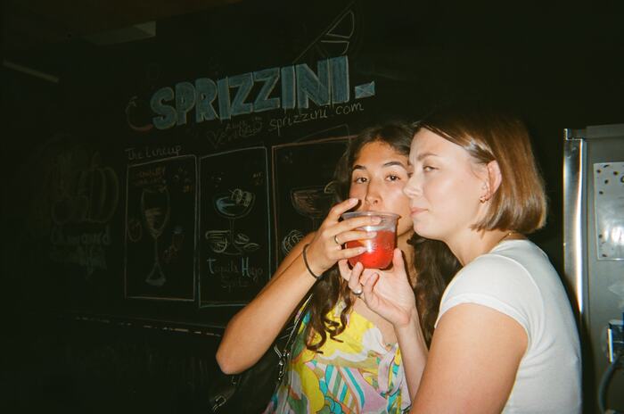 Two guests sharing a red drink in front of the Sprizzini chalkboard