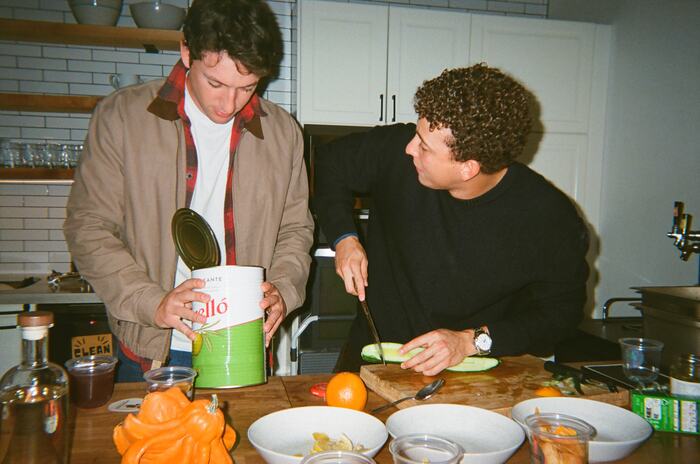 Two guests at the kitchen island, one opening a tin of olives, one slicing cucumber