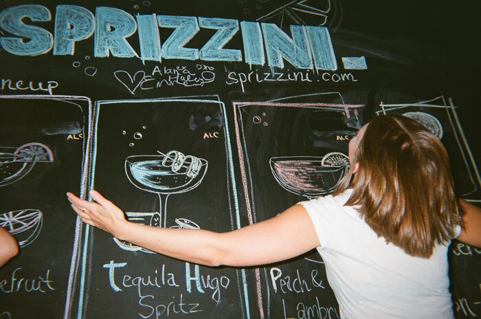 A guest stretching her arms across the Sprizzini chalkboard menu of hand-drawn drinks