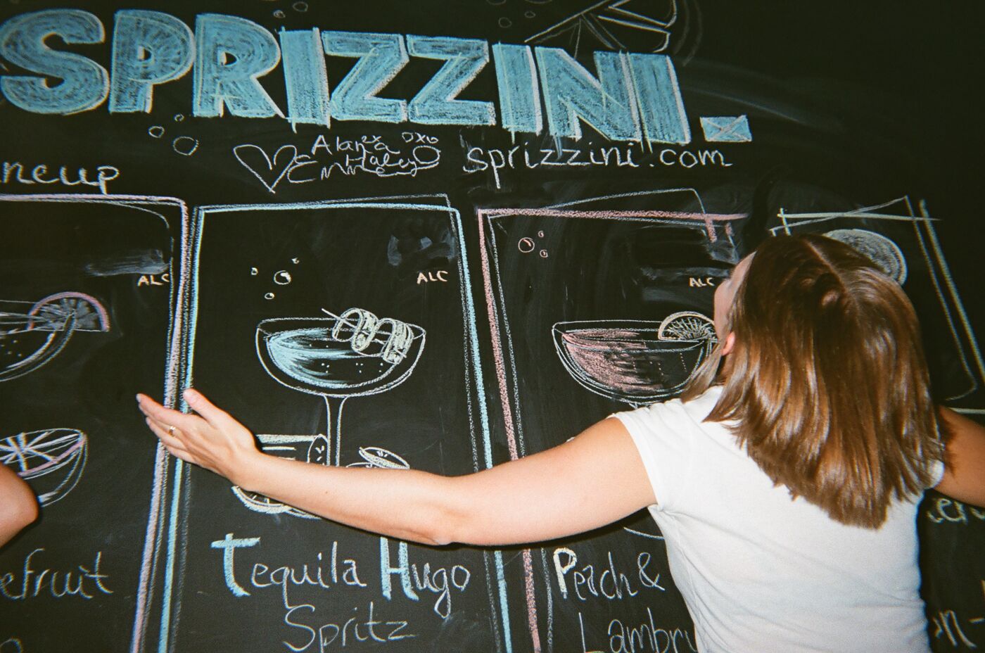 A guest stretching her arms across the Sprizzini chalkboard menu of hand-drawn drinks