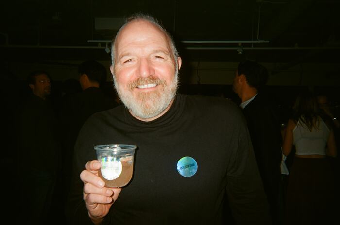 A guest in a black turtleneck beaming, holding up a cup with a Sprizzini sticker