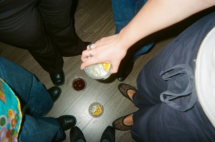 A hand holding a cup over a circle of shoes, two drinks on the floor