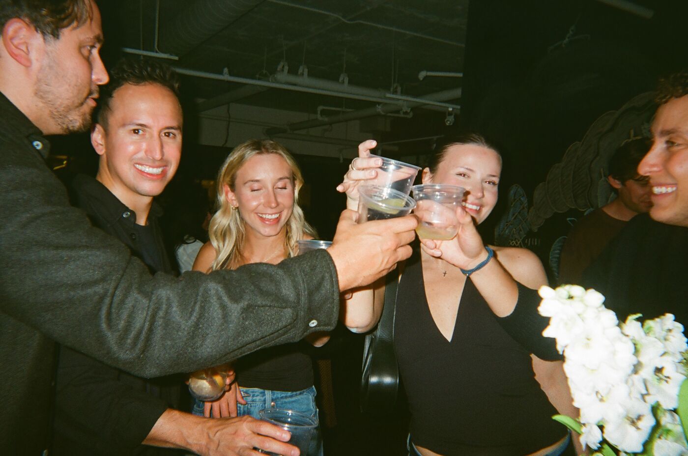 Five guests reaching in to touch cups over a vase of white flowers