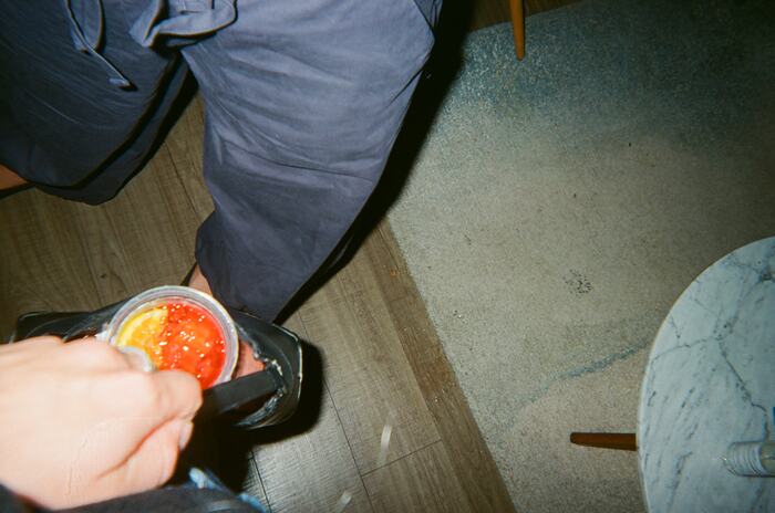 A hand holding a red drink with an orange slice down at knee height