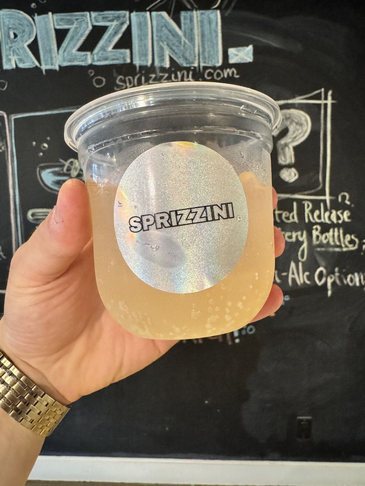 A hand holding a golden spritz in front of the Sprizzini chalkboard