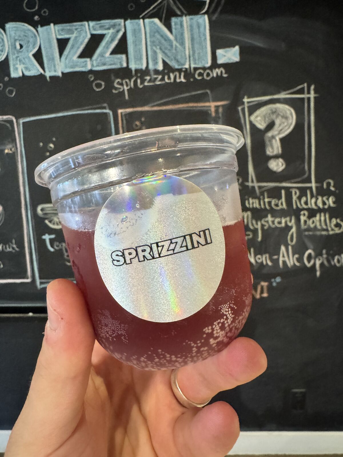 A hand holding a cup of dark red Dirty Picante Negroni in front of the chalkboard menu