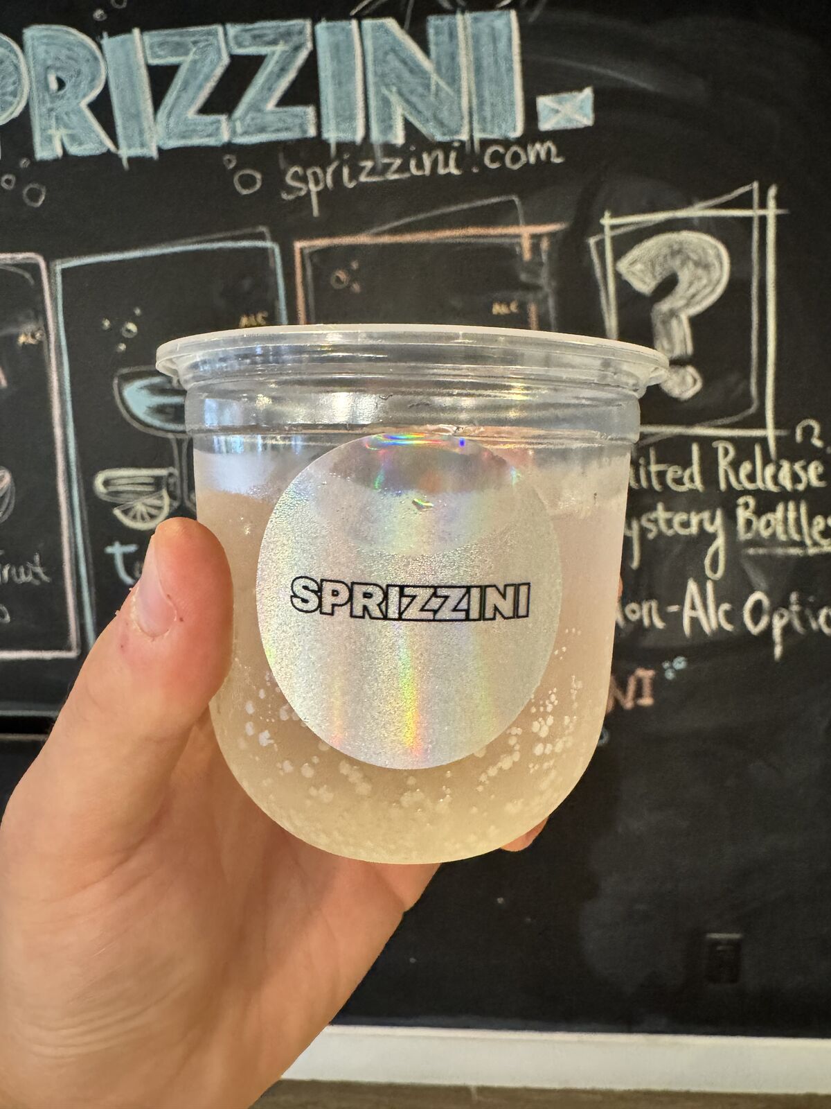 A hand holding a pale spritz in front of the chalkboard listing the limited-release mystery bottles